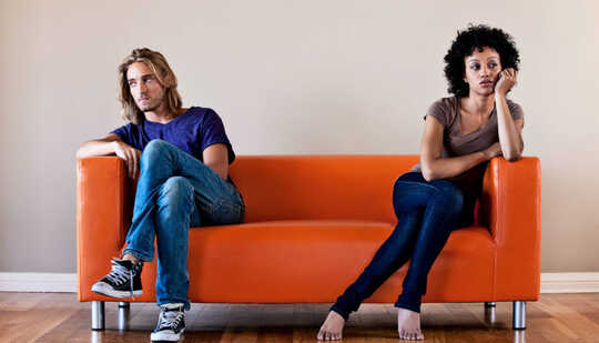 Um casal está sentado em lados opostos de um sofá laranja olhando para longe um do outro