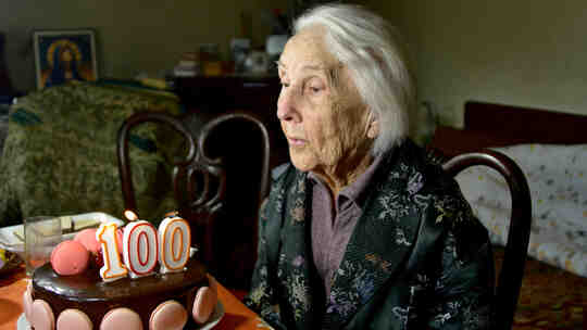 100-letnia kobieta zdmuchuje świeczki na swoim urodzinowym torcie.
