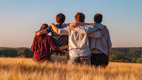 4 orang dewasa muda duduk bersama-sama dengan lengan yang dipaut di bahu mereka - dilihat dari belakang