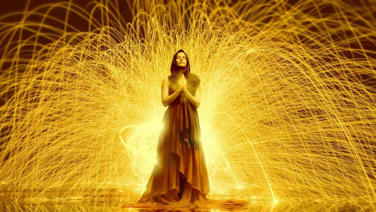 egy nő, kezével ima helyzetben áll, és fénysugarak áramlanak ki belőle