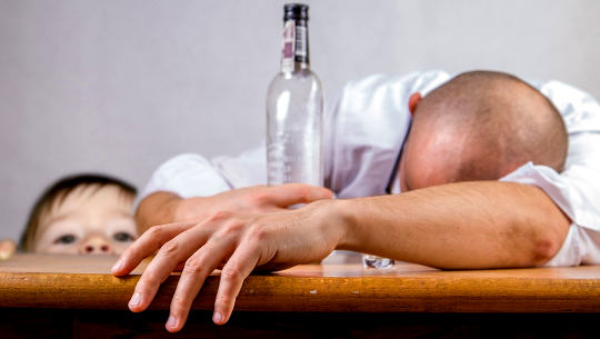 homem desmaiado em uma mesa com uma garrafa vazia de álcool e uma criança olhando
