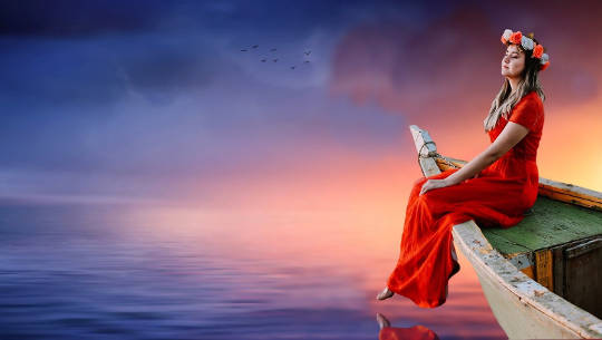 kobieta ubrana w długą sukienkę i girlandę kwiatów na głowie siedzi na brzegu pływającej łodzi o zachodzie słońca