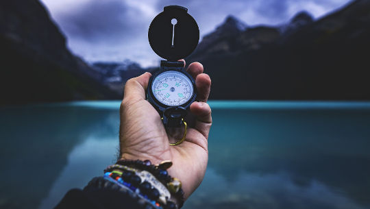 sebuah tangan memegang kompas dengan latar belakang danau dan pegunungan
