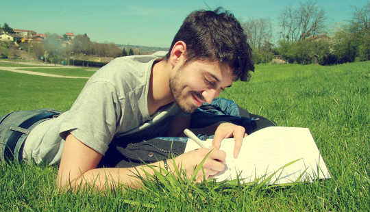 hymyilevä mies makaa nurmikolla kirjoittaen muistikirjaan