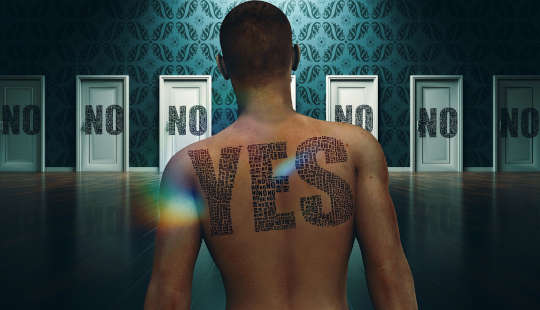 egy férfi, akinek a hátára tetovált IGEN szó van, olyan ajtókkal néz szembe, amelyekre mindegyik NEM-et mond