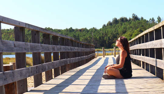 wanita muda menghadap matahari dan duduk di jembatan kayu