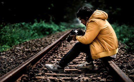người đàn ông trẻ tuổi ngồi trên đường ray nhìn vào hình ảnh trong máy ảnh của mình