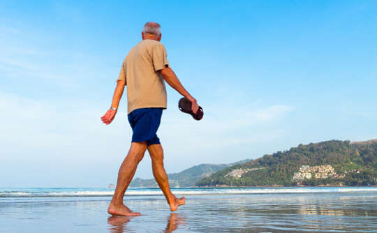 مردی در حال قدم زدن در ساحل در حالی که صندل های خود را در دست گرفته است