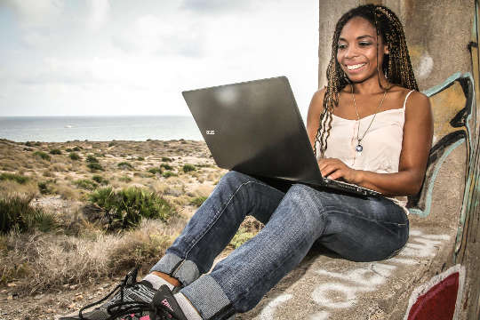 wanita muda duduk dengan punggung bersandar pada pohon bekerja di laptopnya