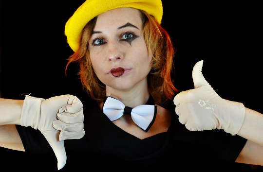 egy csokornyakkendőt és bohóc arcot viselő fiatal nő fehér kesztyűvel, felfelé és lefelé hüvelykujjjal