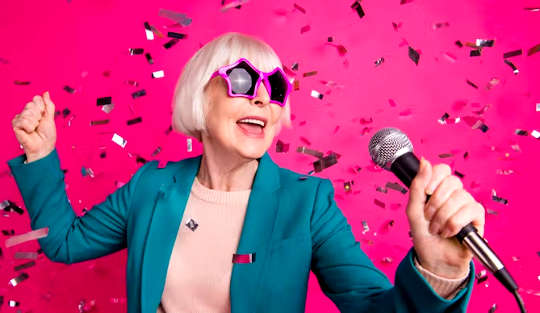 마이크를 잡고 노래하는 펑키한 분홍색 선글라스를 쓴 회색 머리 여자