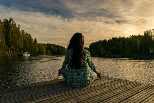 woman, seen from the back, sitting in a lotus pose on a dock by a lake