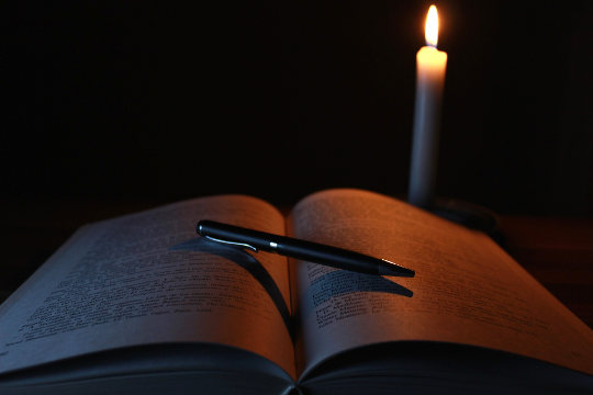 egy nyitott könyv, rajta egy toll és egy gyertya világít a könyvön