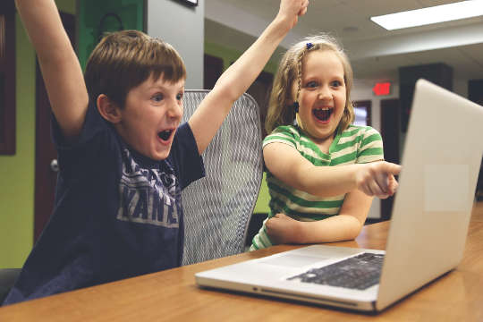 двое детей перед компьютером празднуют успех, поднимают руки вверх и широко улыбаются