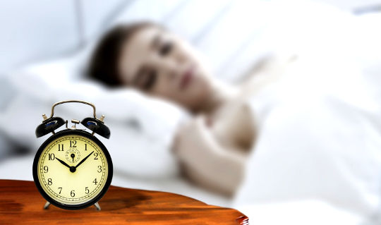 ایک عورت رات کے اسٹینڈ پر پرانے طرز کی غیر الیکٹرانک الارم گھڑی کے ساتھ سو رہی ہے۔