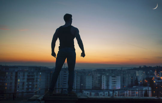 uma silhueta de um homem com os punhos cerrados em pé em um telhado com vista para a cidade