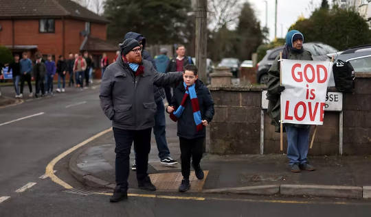 φίλαθλοι στο δρόμο για έναν αγώνα, περαστικός που κρατά μια πινακίδα GOD IS LOVE