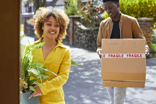 femme tenant une plante en pot, homme tenant une boîte qui dit Fragile, entrant dans une maison