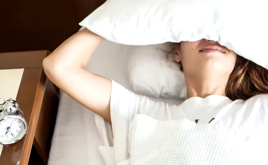 pessoa deitada na cama com travesseiro na cabeça e despertador ao lado da cama