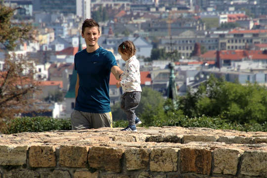ett litet barn som går på toppen av en stenmur med pappa stående genom att le och hålla barnets hand