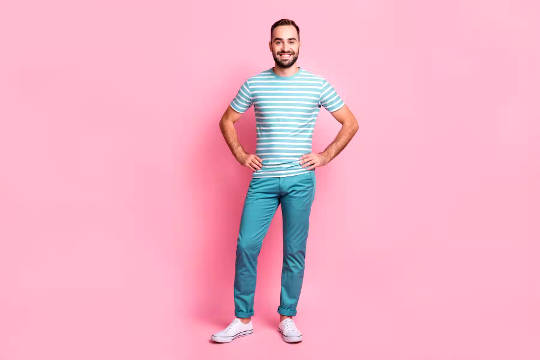 un hombre parado frente a una pared rosa con las manos en las caderas y una gran sonrisa