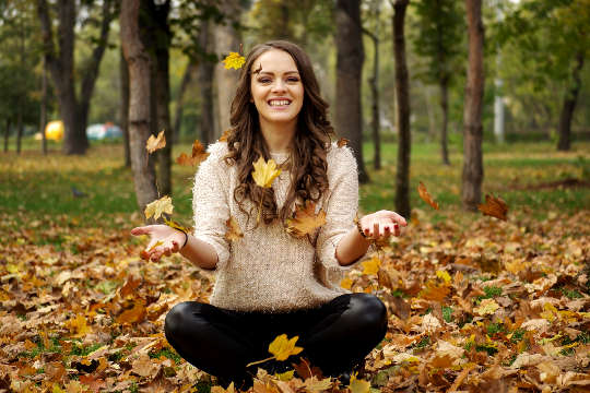 une jeune femme souriante assise dans la forêt au milieu des feuilles d'automne qui tombent
