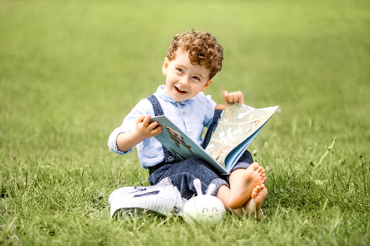 menino sorridente sentado do lado de fora com um livro aberto nas mãos