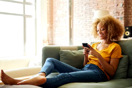 una giovane donna nera seduta rilassata sul suo divano usando il suo telefono