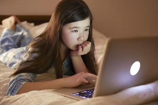 एक युवा लड़की अपने बिस्तर पर लेटी हुई एक वेबकैम की नज़र के नीचे लैपटॉप का उपयोग कर रही है