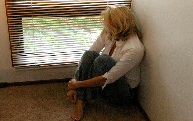 Frau sitzt auf dem Boden und blickt durch die Ritzen einer Jalousie eines Fensters