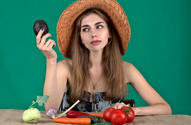 egy nő, egy sor friss zöldséggel maga előtt, és egy avokádót tart fel