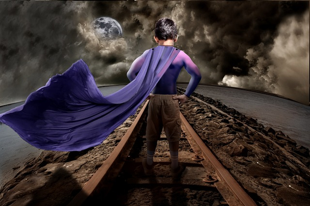 young boy standing on a railroad track and wearing a superhero cape