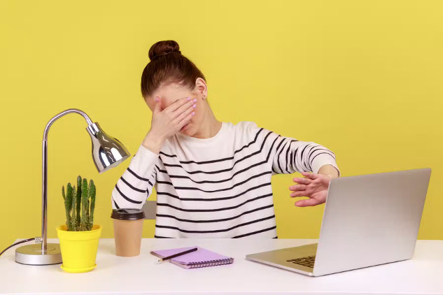 A young woman sitting at a desk in front of a yellow wall, puts one hand over her eyes and uses the other to shield her computer screen, suggesting 'I do not want to look at this'.