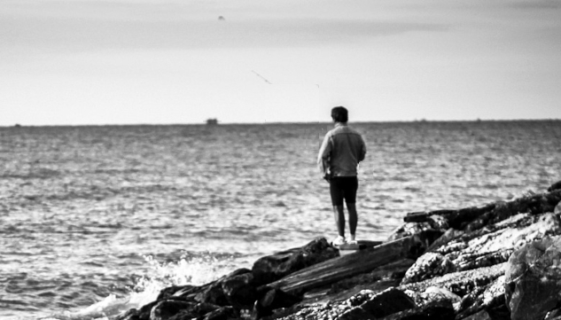 Image of a man standing on some rocks looking over at the water. Image by Davide Dalese from Pixabay. 