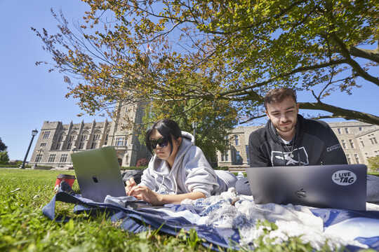 Twee studenten zitten in het gras met laptops die naast elkaar in de buitenlucht studeren.