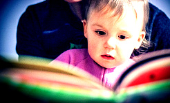 एक बच्चा अपनी माँ की गोद में बैठा है और किताब पढ़ रहा है