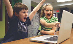 dwoje dzieci przed komputerem świętuje sukces z rękami w górze i dużymi smielami