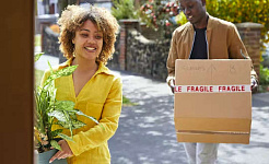 femeie ținând o plantă în ghiveci, bărbat ținând o cutie pe care scrie Fragil, intrând într-o casă