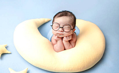 baby with eyes closed wearing huge glasses and resting on a crescent moon-shaped pillos
