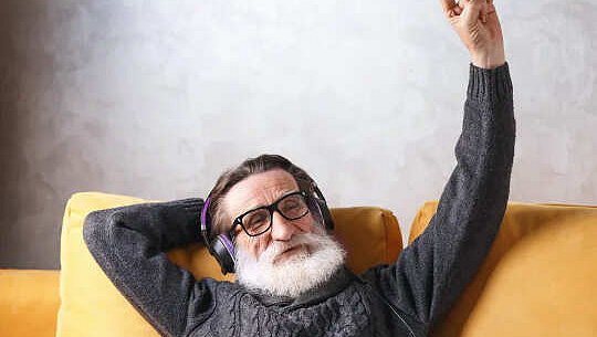 Homme à la barbe blanche, portant des écouteurs, assis sur un canapé et faisant un signe Shaka avec sa main gauche.