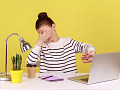 一名年輕女子坐在黃牆前的桌子旁，一隻手摀住眼睛，另一隻手擋住電腦螢幕，暗示「我不想看這個」。
