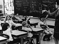 A white woman stands by a classroom blackboard in front of white students sitting at desks, many with their hands raised.