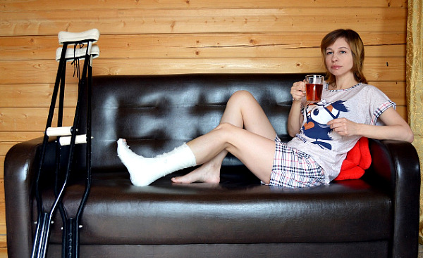 wanita duduk di sofa sambil minum teh dengan kaki digips dan kruk di sisinya