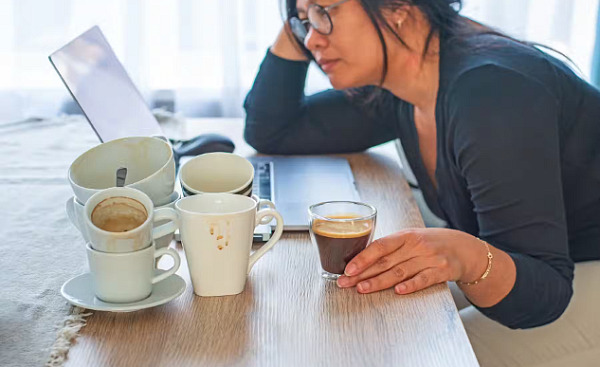 kobieta wyglądająca na zestresowaną i zmęczoną, pijąca filiżankę kawy i otoczona wieloma pustymi i pełnymi filiżankami