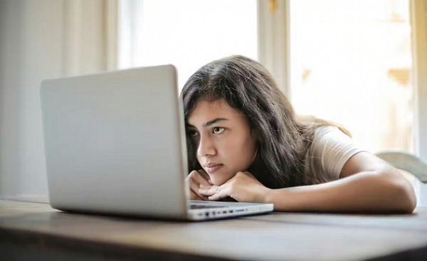 A teenage girl leans her head on a table and looks at a laptop screen.