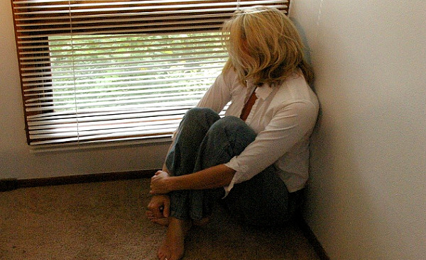 nainen istuu lattialla ja katsoo ulos ikkunan sälekaihtimien halkeamista
