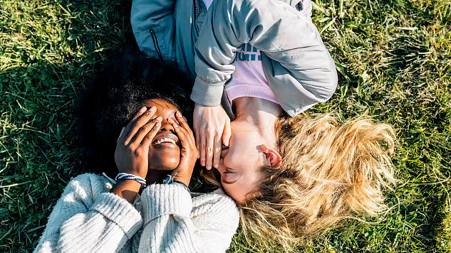 Two smiling people lying on grass, one with hands over eyes and the other whispering into their ear.