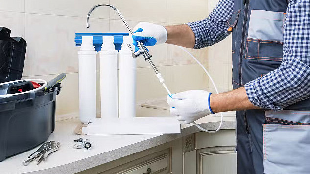 A man in work clothes installing a water filtration system under the kitchen sink, representing the process of reducing PFAS contamination in household water.