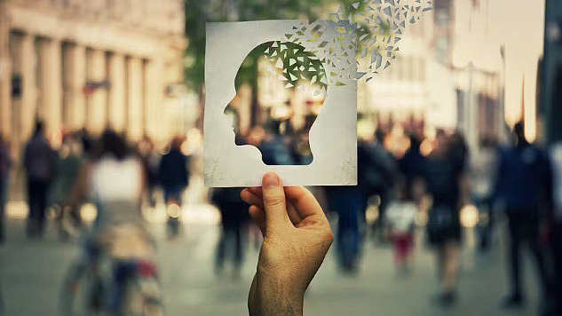 Hand holding a paper sheet with a human head icon broken into pieces over a crowded street background.