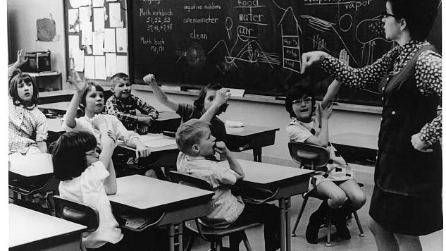 A white woman stands by a classroom blackboard in front of white students sitting at desks, many with their hands raised.